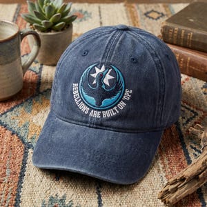 May include: A navy blue baseball cap with a circular emblem featuring a bird and star design in white and teal. The text "REBELLIONS ARE BUILT ON OPE" is embroidered around the emblem. The cap is made of a soft, worn fabric.