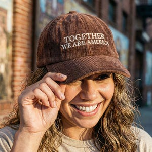 Może przedstawiać: Brązowa czapka bejsbolowa sztruksowa z białym napisem "TOGETHER WE ARE AMERICA". Czapka ma zakrzywiony daszek i zaokrągloną koronę. Czapka jest noszona przez osobę.