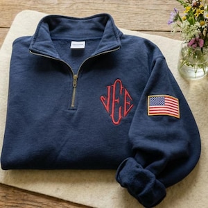 Könnte beinhalten: Marineblaues Sweatshirt mit Viertelreißverschluss, rotem Monogramm und einem amerikanischen Flaggenaufnäher auf dem Ärmel. Das Sweatshirt hat einen gerippten Bund und einen Messingfarbenen Reißverschluss.