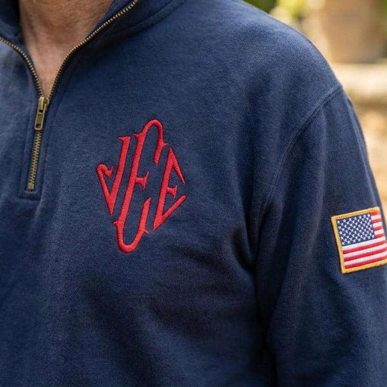 May include: Navy blue sweatshirt with a quarter-zip neckline and a red embroidered design on the chest. The sweatshirt features a small American flag patch on the sleeve.
