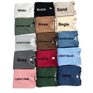May include: A collection of crewneck sweatshirts in various colors, including white, black, sand, military green, brown, sports grey, dark chocolate, grey, maroon, light pink, forest green, and navy. Each sweatshirt has a label with its color name.