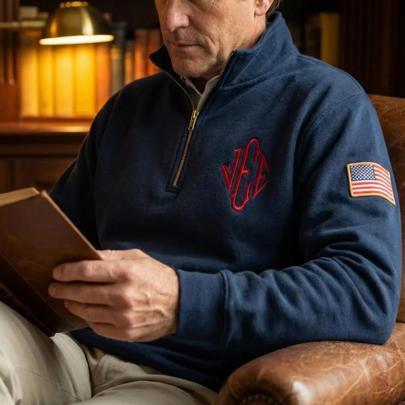 May include: Navy blue quarter-zip sweatshirt with a red embroidered logo and an American flag patch on the sleeve. The sweatshirt has a gold zipper and a high collar. The person is holding a book.