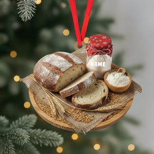 May include: A round, wooden Christmas ornament featuring a loaf of sliced bread, wheat, flour, and a jar with the text "NAME". The ornament has a red ribbon for hanging. The bread is brown with a white dusting.