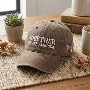 May include: A brown baseball cap with the text "TOGETHER WE ARE AMERICA" embroidered in white. The cap features a small American flag patch on the side. The hat is sitting on a woven mat.