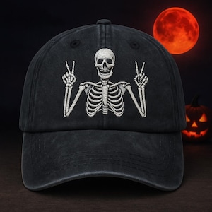 May include: Black baseball cap with a white embroidered skeleton giving the peace sign. The cap has a curved peak and a button on top. A red full moon and a carved pumpkin are in the background.