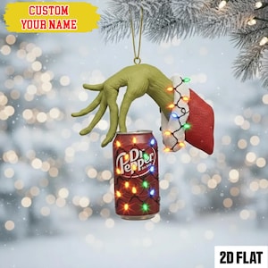 May include: A Christmas tree ornament depicting a green hand reaching for a Dr. Pepper can, decorated with fairy lights. The hand wears a Santa Claus cuff. The ornament has a gold hanging string and the text "CUSTOM YOUR NAME".