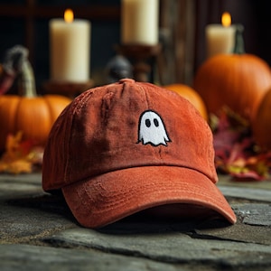 May include: A rust-coloured baseball cap featuring a white ghost embroidery. The hat is made of a soft, textured material. Pumpkins and candles are in the background, suggesting a Halloween theme.