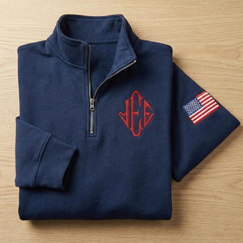 May include: Navy blue quarter-zip sweatshirt with a red monogram and an American flag patch on the sleeve. The sweatshirt has a ribbed collar and cuffs, and a silver zipper. The monogram is in a diamond shape.