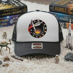 May include: A black and white trucker hat with a gray brim and a white front panel. The front features a circular design with the text "PROJECT HAIL MARY" and an illustration of a spaceship. The hat has a mesh back.