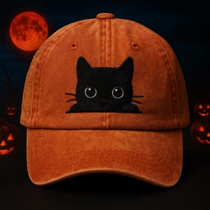 Può includere: Cappellino da baseball arancione con un gatto nero ricamato sul davanti. Il gatto ha occhi e baffi bianchi. Lo sfondo include una luna piena arancione e zucche di Halloween, creando un tema di Halloween.