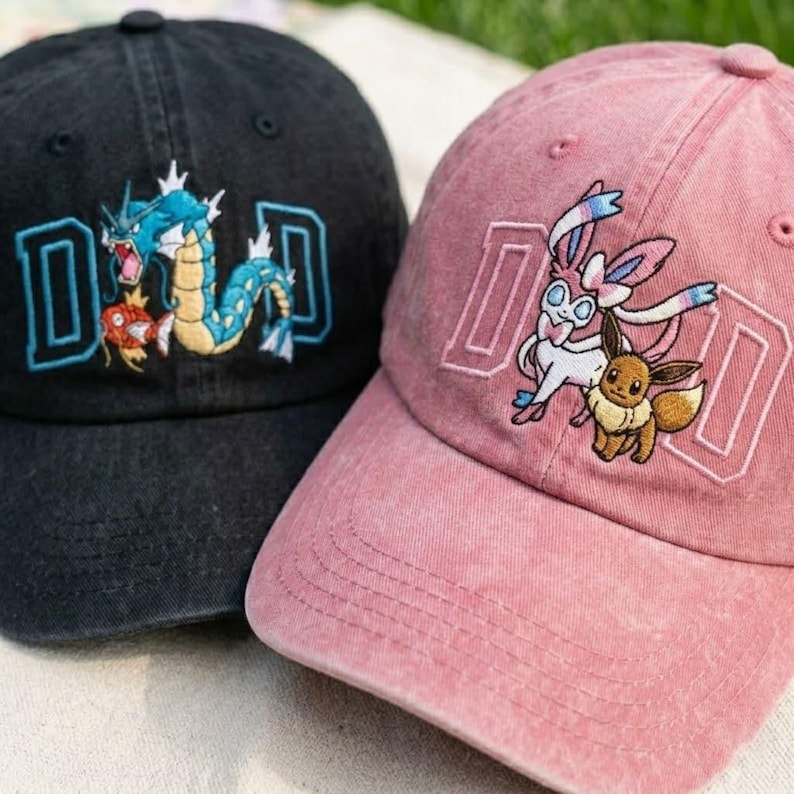 May include: Two baseball caps, one black and one pink, with embroidered designs. The black cap features a blue and orange dragon-like creature with the letter "D" in a blue outline. The pink cap has two cartoon characters with the letter "D".