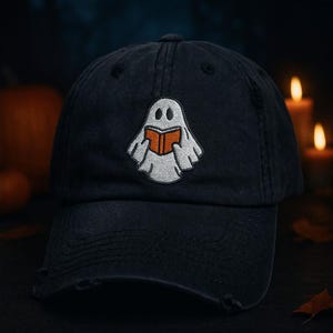 May include: A navy blue baseball cap with a distressed brim. The cap features an embroidered ghost reading a book with an orange cover. The ghost is white with black eyes and is holding the book with its hands. The background is dark.