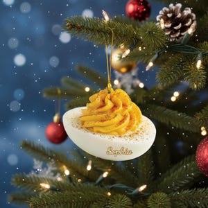 Könnte beinhalten: Ein Weihnachtsbaumschmuck in Form eines gefüllten Eies, der an einem geschmückten Baum hängt. Der Schmuck ist weiß mit gelber Füllung und Paprikastaub. Der Name "Sophia" ist auf dem Schmuck geschrieben.