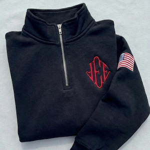 May include: Black quarter-zip sweatshirt with a silver zipper and a red embroidered monogram. The sweatshirt features an American flag patch on the upper left sleeve. The fabric appears soft and comfortable.