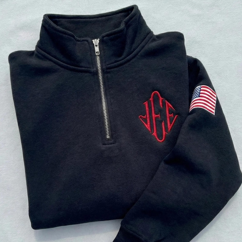 May include: Black quarter-zip sweatshirt with a silver zipper and a red embroidered monogram. The sweatshirt features an American flag patch on the upper left sleeve. The fabric appears soft and comfortable.