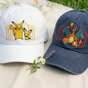 May include: Two baseball caps with embroidered designs. One is white with a yellow Pikachu and Pichu design, and the other is navy blue with a Charizard and Charmander design. Both caps have the word "DAD" embroidered in a matching color.
