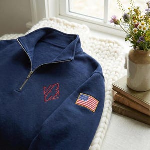 May include: Navy blue quarter-zip sweatshirt with a red embroidered logo and an American flag patch on the sleeve. The sweatshirt has a ribbed collar and a silver zipper. The garment is made of a soft, textured fabric.