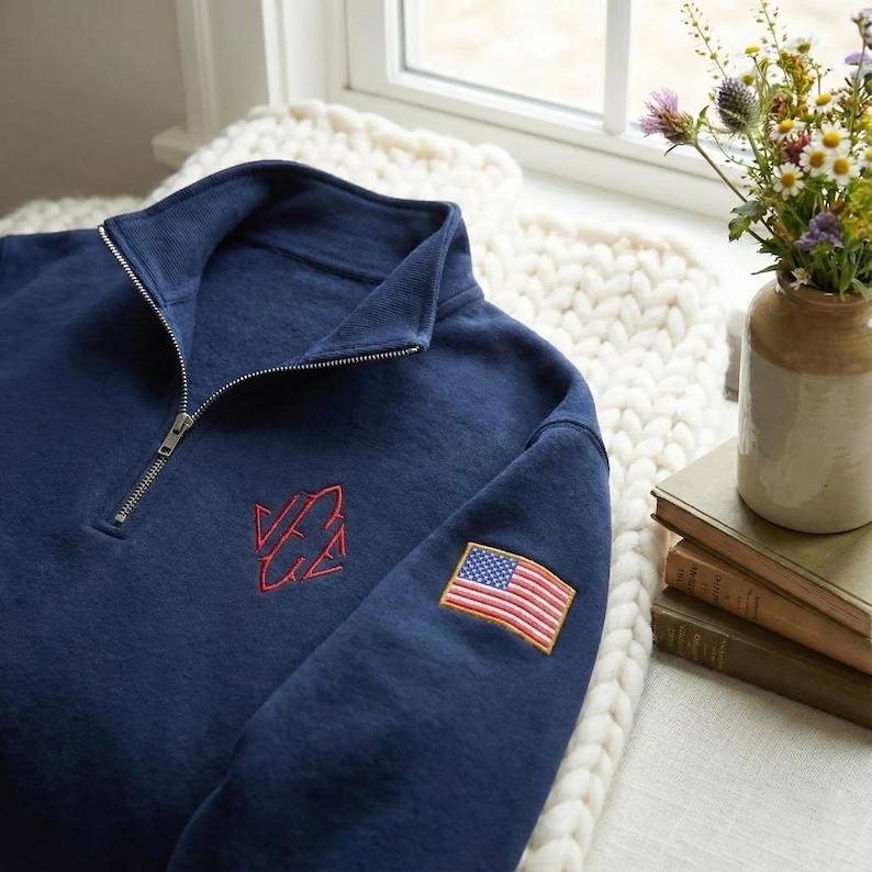 May include: Navy blue quarter-zip sweatshirt with a red embroidered logo and an American flag patch on the sleeve. The sweatshirt has a ribbed collar and a silver zipper. The garment is made of a soft, textured fabric.