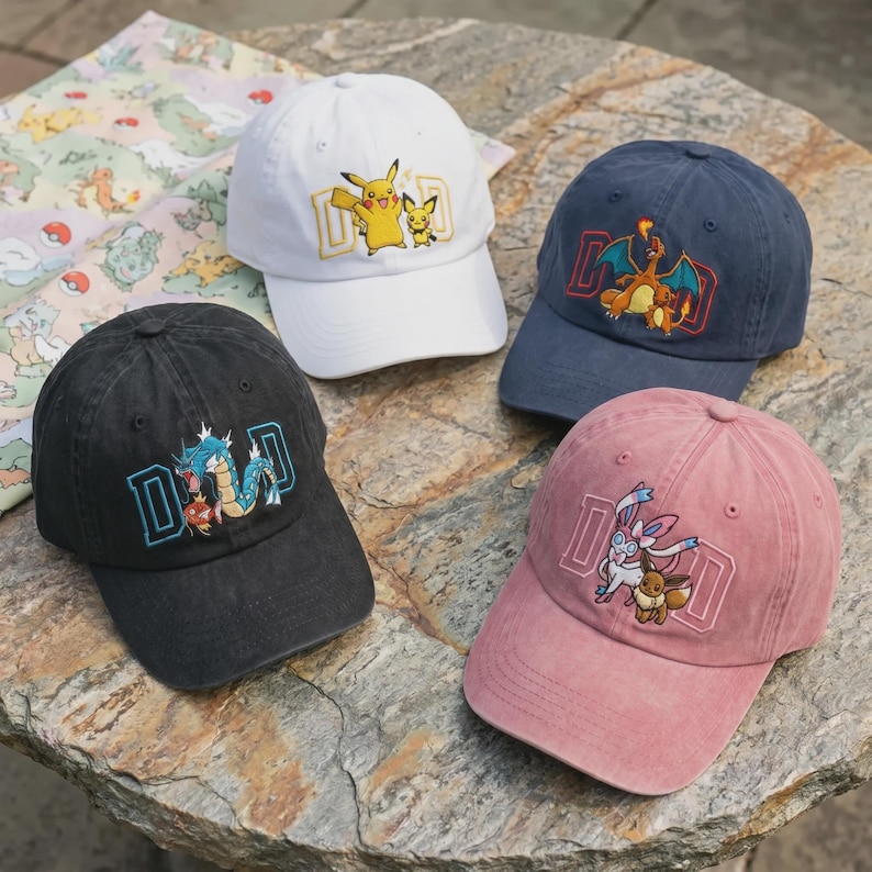 May include: Four baseball caps in white, navy blue, black, and pink. Each cap features a different Pokemon character design within a rectangular frame. The caps are displayed on a stone surface.