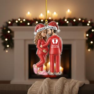 Op de afbeelding: Een rode kerstversiering met een stel in bijpassende pyjama's en kerstmutsen. De vrouw is zwanger en de man heeft zijn arm om haar heen. De ornamenten hebben de namen "Sophia" en "Thomas" en het jaar "2026".