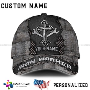 Puede incluir: Una gorra de béisbol personalizada con un diseño de metal gris y negro. La gorra presenta una cruz con dos llaves inglesas, el texto "Iron Worker" y un espacio para agregar un nombre personalizado.