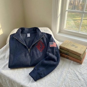 May include: Navy blue quarter-zip sweatshirt with a red embroidered monogram and an American flag patch on the sleeve. The sweatshirt is folded on a white surface, with a window and books in the background.