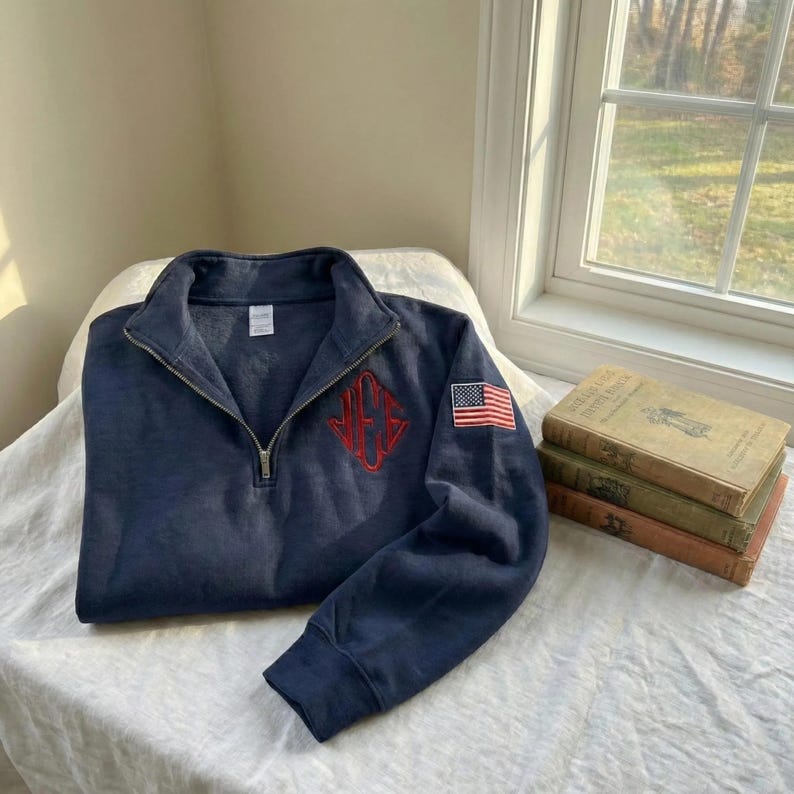 May include: Navy blue quarter-zip sweatshirt with a red embroidered monogram and an American flag patch on the sleeve. The sweatshirt is folded on a white surface, with a window and books in the background.