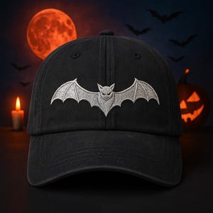 Könnte beinhalten: Schwarze Baseballkappe mit einem silbernen, gestickten Fledermaus-Design. Die Fledermaus hat ausgebreitete Flügel und ein detailliertes Gesicht. Die Kappe hat ein klassisches Sechs-Panel-Design, ideal für Halloween.