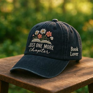 Może przedstawiać: Granatowa czapka bejsbolówka z haftowanym napisem "Just One More Chapter" i "Book Lover". Czapka ma kwiatowy wzór z białymi i różowymi kwiatami nad otwartą książką. Kapelusz leży na drewnianej powierzchni.