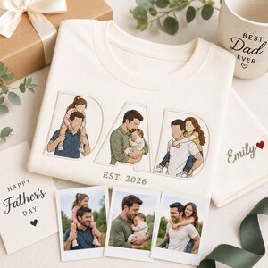 Puede incluir: Sudadera color crema con la palabra "DAD" bordada en letras grandes, con ilustraciones de un padre con sus hijos. La sudadera tiene "EST. 2026" bordado debajo. Incluye una taza, una tarjeta y fotos, todo con tem&aacute;tica del D&iacute;a del Padre.