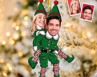 May include: A Christmas ornament featuring a couple dressed as elves. The ornament has a green and red color scheme, with the couple's faces visible. The elf costumes include hats, collars, and striped socks. A gold string is attached for hanging.