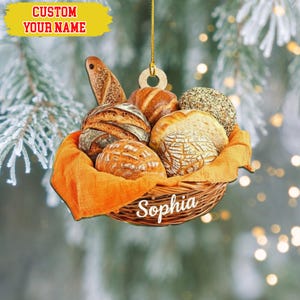 May include: A wooden Christmas ornament shaped like a basket filled with various types of bread. The basket is lined with orange fabric and has the name "Sophia" written on it. The ornament also has the text "CUSTOM YOUR NAME".