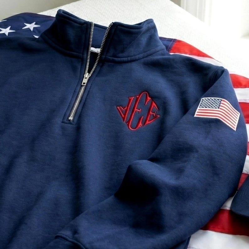 May include: Navy blue quarter-zip sweatshirt with a red embroidered logo and an American flag patch on the sleeve. The sweatshirt has a silver zipper and is made of a soft material.