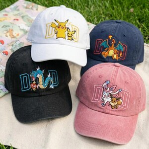 May include: Four baseball caps in white, navy blue, black, and pink. Each cap features a different embroidered cartoon character design with the letters "DAD" in a block letter style. The caps are on a green grassy surface.