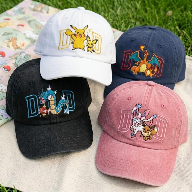 May include: Four baseball caps in white, navy blue, black, and pink. Each cap features a different embroidered cartoon character design with the letters "DAD" in a block letter style. The caps are on a green grassy surface.