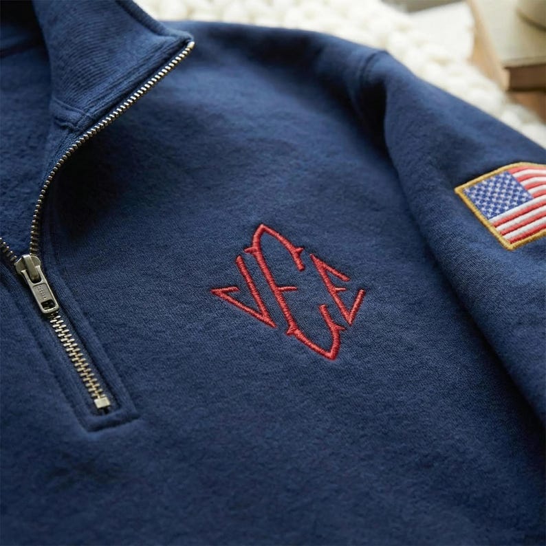 May include: Navy blue quarter-zip sweatshirt with a red embroidered monogram and an American flag patch on the sleeve. The sweatshirt features a ribbed collar and a metal zipper. The fabric appears to be a soft, textured material.