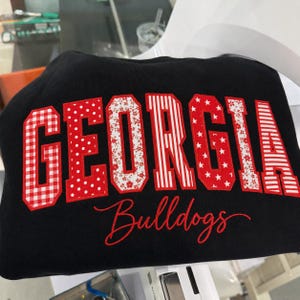Può includere: Una felpa nera con la parola GEORGIA in grandi lettere stampatello. Ogni lettera presenta un motivo unico in rosso e bianco, tra cui quadretti vichy, pois, fiori, righe e stelle. La parola Bulldogs è ricamata in corsivo rosso sotto.