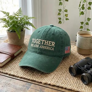 May include: A green baseball cap with the text "TOGETHER WE ARE AMERICA" embroidered in white. The cap features a small American flag patch on the side. The hat is sitting on a woven mat, with a small potted plant and a pair of binoculars in the background.