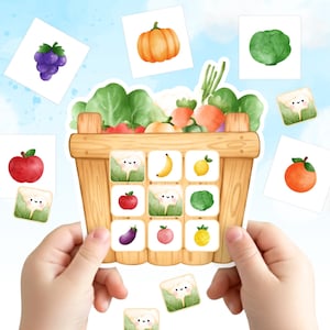 May include: A wooden basket filled with watercolor illustrations of vegetables and fruits, including carrots, cauliflower, and a pumpkin. The basket is held by two hands, with additional fruit and vegetable cards scattered around.