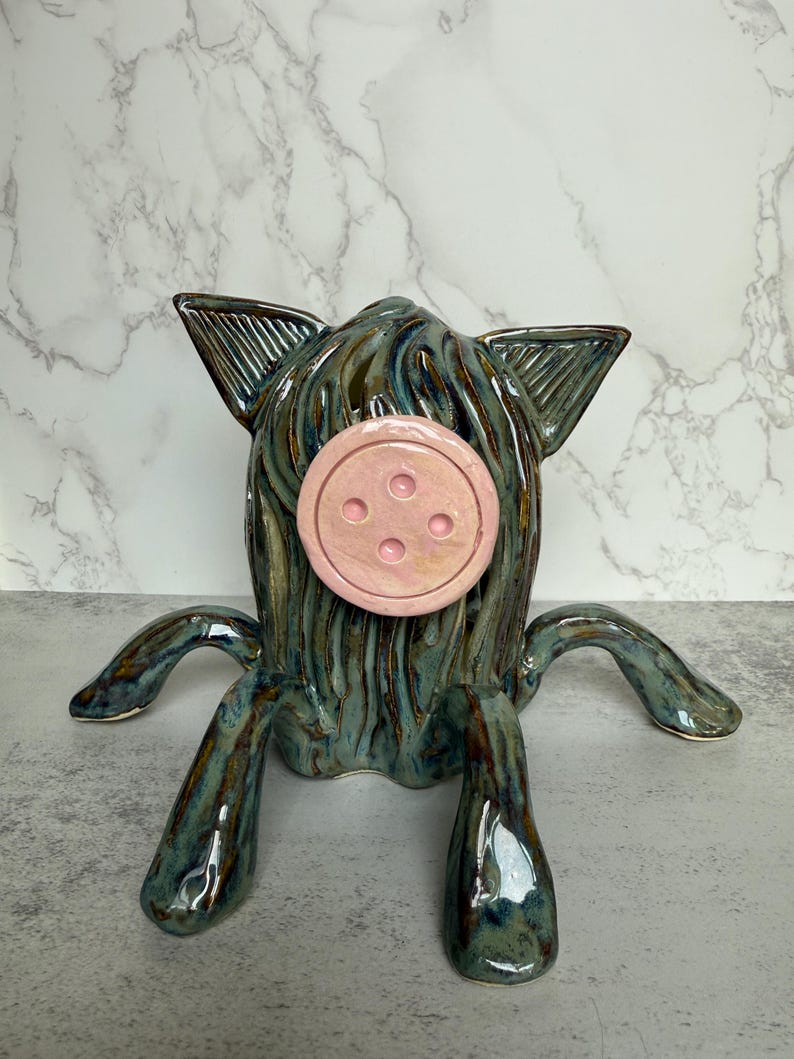 May include: A whimsical ceramic sculpture of a creature with a teal and brown glaze. It features a pink, button-shaped nose and four legs. The ears are pointed and the overall design is unique and artistic.