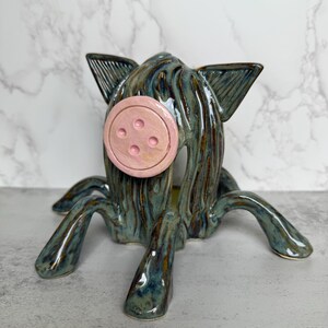May include: A ceramic pig sculpture with a button nose and four legs. The pig is glazed in shades of teal and brown, with a pink button nose. The sculpture has a whimsical, artistic design.