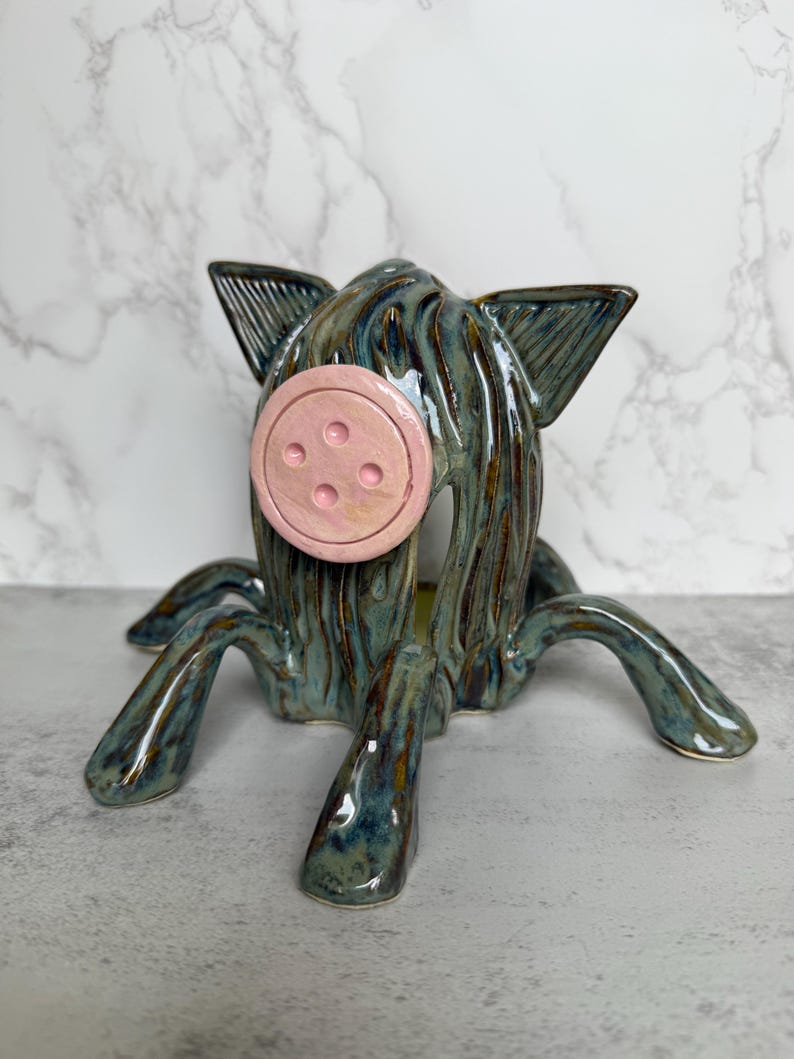 May include: A ceramic pig sculpture with a button nose and four legs. The pig is glazed in shades of teal and brown, with a pink button nose. The sculpture has a whimsical, artistic design.