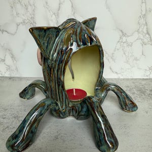 May include: A ceramic teal and brown cat-shaped candle holder with a red candle inside. The candle holder has a textured surface and four legs. The interior is a light yellow color.