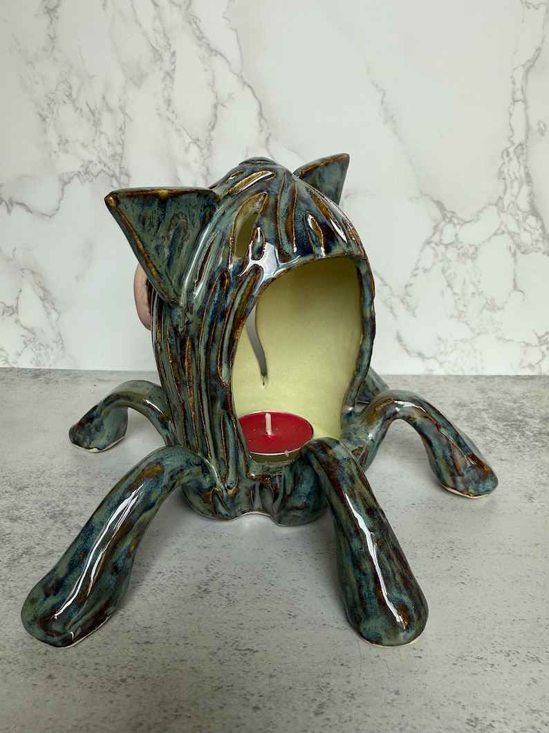 May include: A ceramic teal and brown cat-shaped candle holder with a red candle inside. The candle holder has a textured surface and four legs. The interior is a light yellow color.