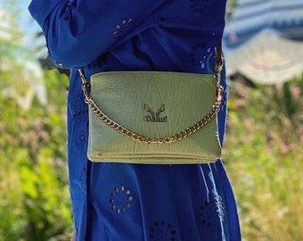 Könnte beinhalten: Eine Frau trägt ein blaues Kleid mit weißen Stickereien und eine hellgrüne Umhängetasche mit einer goldenen Kettenkette. Die Tasche hat ein kleines, geprägtes Logo auf der Vorderseite.