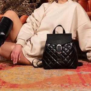 May include: A woman wearing a cream coloured sweater and black knee-high boots sits on a red velvet sofa with a black quilted backpack in front of her. A leopard print jacket is draped over the back of the sofa.