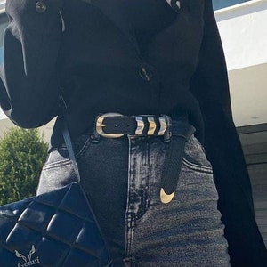 May include: A woman wearing a black blazer, black top, and dark wash jeans with a silver belt. She is carrying a black quilted shoulder bag with a silver logo that says "Genuf."