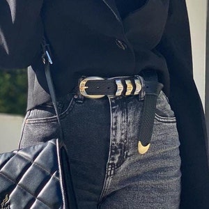 May include: A black quilted shoulder bag with a gold metal logo that says "Genny". The bag is worn over a black blazer and dark wash denim jeans with a gold and black belt.
