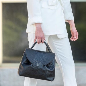 May include: A woman wearing a white blazer with a matching white pantsuit and a black leather handbag with the word "Gemini" embossed on the front.