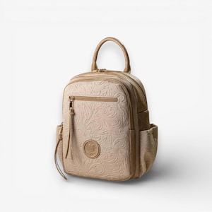 Puede incluir: Mochila beige con un patrón floral en relieve. La bolsa tiene un asa superior, un bolsillo frontal con cremallera y bolsillos laterales. Un pequeño emblema redondo está centrado en la parte delantera. Los herrajes son dorados.